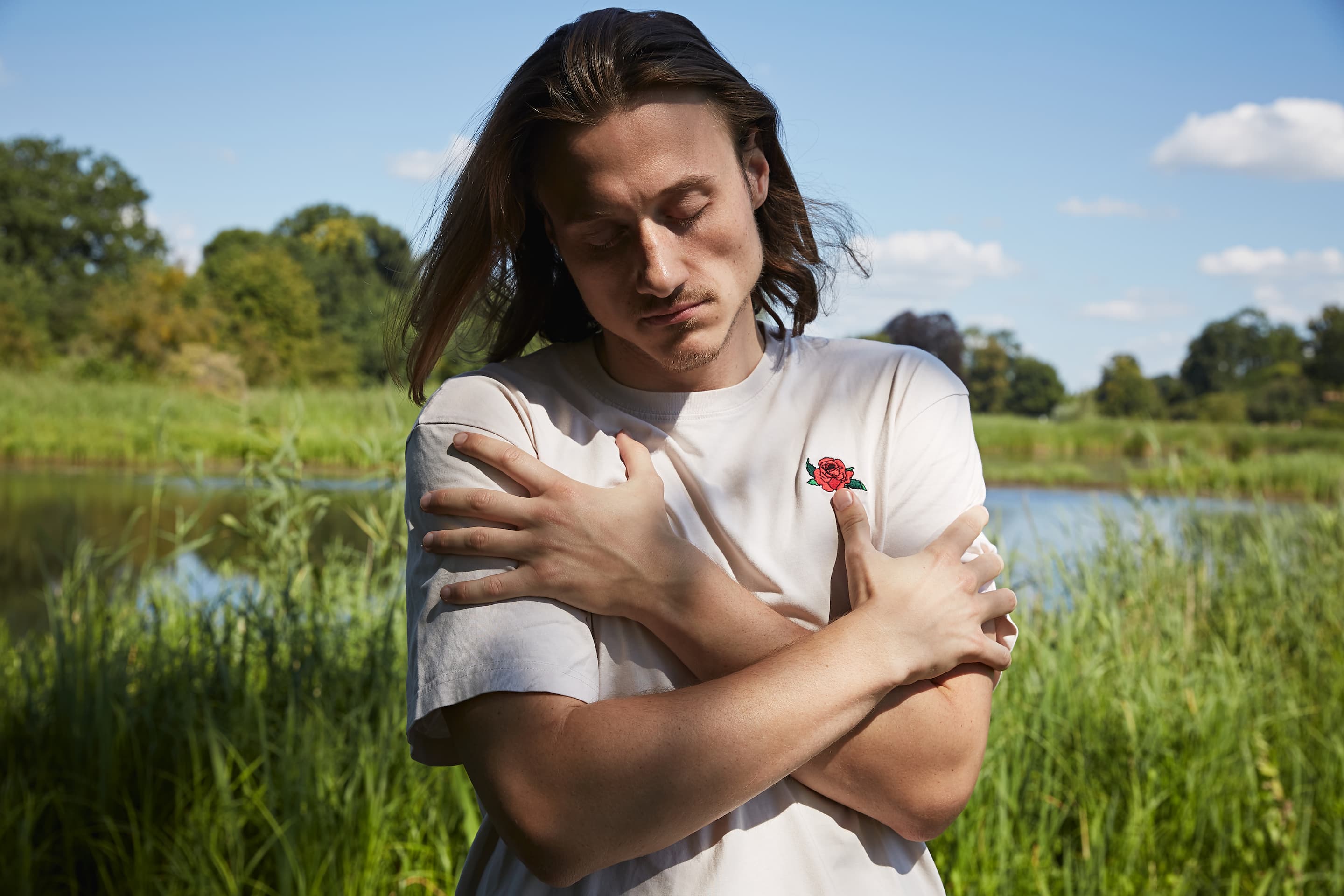 Eine Person mit langen Haaren steht im Freien an einem Gewässer und hat die Arme vor der Brust verschränkt. Sie trägt ein weißes T-Shirt mit einem kleinen roten Rosenmuster darauf. Der Hintergrund ist grün und sonnig.