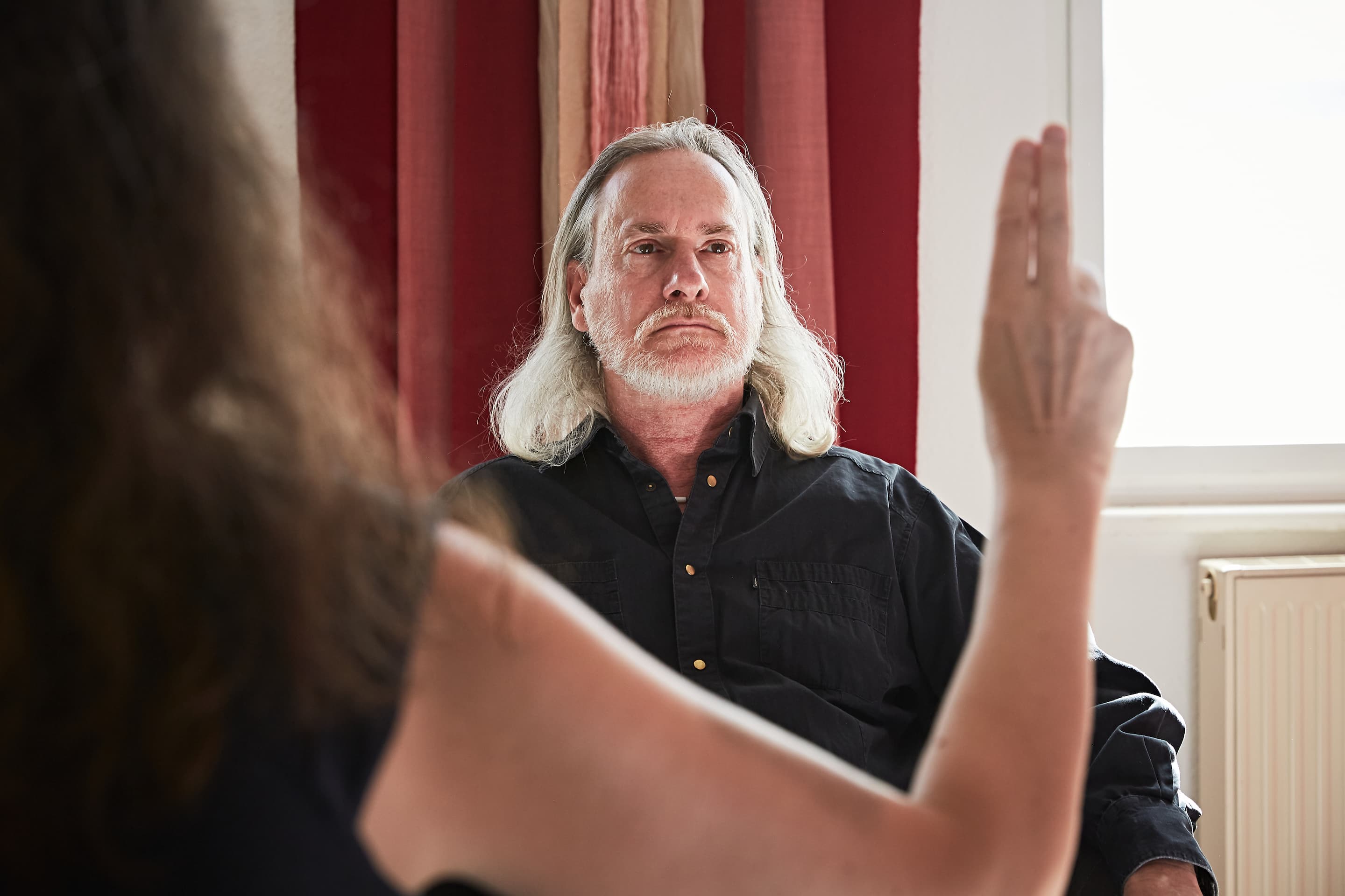 Ein Mann mit langen grauen Haaren sitzt und blickt eine Person an, die zwei Finger vor sich hochhält.
