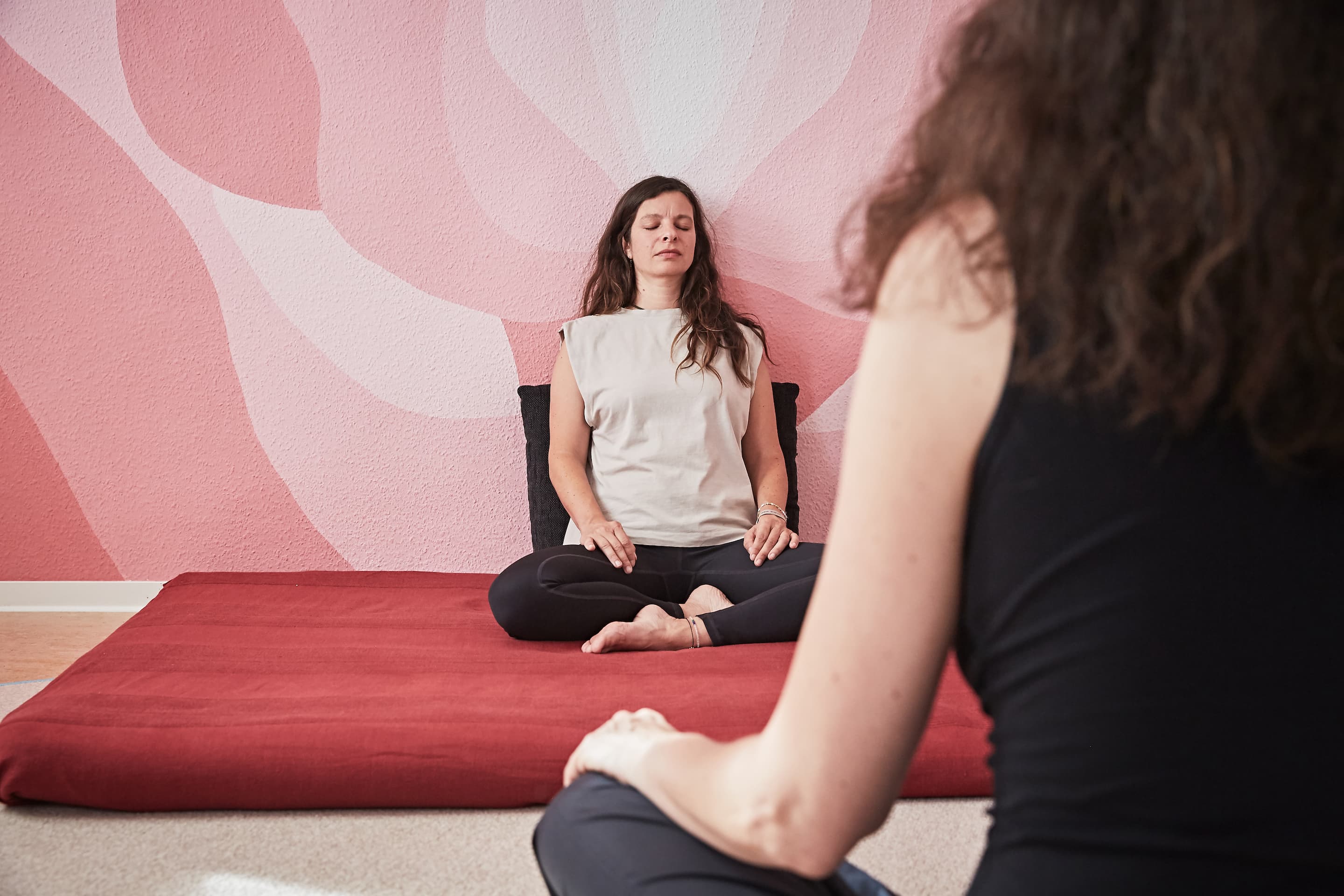 Zwei Frauen in einem friedlichen Raum während einer Session. Eine sitzt auf einem roten Kissen vor einer rosa Wand und hat die Augen geschlossen, die andere sitzt etwas weiter vorne im Bild.